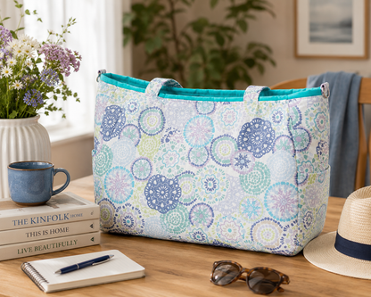 Patterned tote bag on a table with books, mug, notebook, pen, sunglasses, and hat.